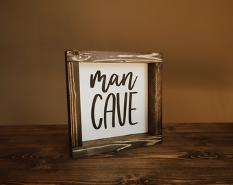 Puede incluir: Un letrero de madera con las palabras "man cave" en letras negras sobre un fondo blanco. El letrero está enmarcado en un marco de madera oscura.