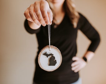 Puede incluir: Un adorno de madera con forma del estado de Michigan, pintado de negro sobre un fondo de madera blanca. El adorno está colgado de una cuerda blanca.