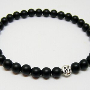 May include: Black beaded bracelet with a silver charm featuring the letter "M". The bracelet is made of small, round, matte black beads strung together. The charm is a small, silver-colored metal piece.