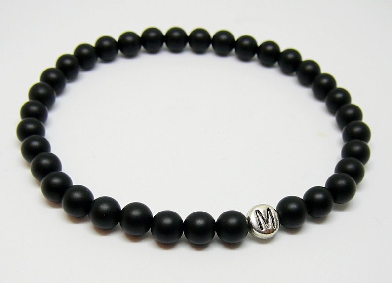 May include: Black beaded bracelet with a silver charm featuring the letter "M". The bracelet is made of small, round, matte black beads strung together. The charm is a small, silver-colored metal piece.