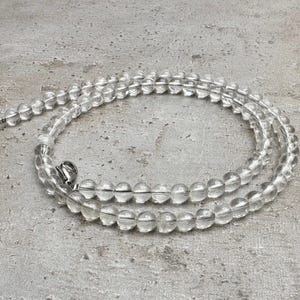 Op de afbeelding: Een ketting van heldere kwarts kralen met een zilveren sluiting. De ketting bestaat uit ronde, transparante kralen die aan elkaar zijn geregen. De kralen zijn gelijkmatig van grootte en creëren een delicate, elegante uitstraling. De ketting ligt op een getextureerd grijs oppervlak.