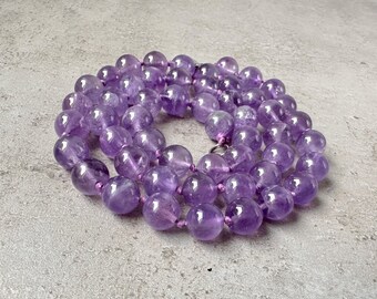 May include: A long strand of amethyst beads, made of round, polished stones. The beads are a light purple color and are strung on a clear thread.