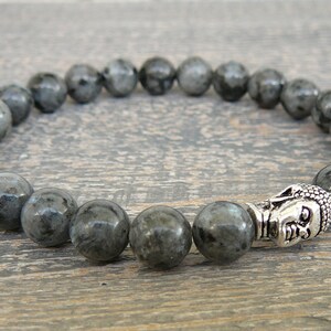 May include: A gray beaded bracelet with a silver Buddha head charm. The beads are round and have a speckled appearance.
