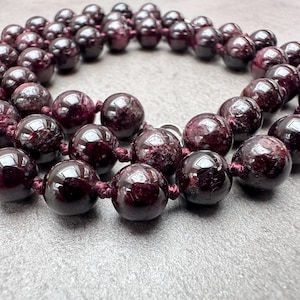 Könnte beinhalten: Eine Halskette aus zahlreichen, tiefroten, kugelförmigen Perlen. Die Perlen sind mit einer passenden roten Kordel aufgefädelt, wodurch eine durchgehende Schlaufe entsteht. Die polierten Perlen reflektieren das Licht und unterstreichen ihre satte Farbe und glatte Textur. Die Halskette ist ein Schmuckstück.