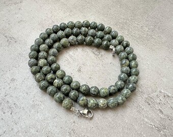 Puede incluir: Una hebra de cuentas de serpentina verde con un cierre plateado. Las cuentas son redondas y tienen un patrón moteado.