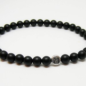 May include: Black beaded bracelet with a silver charm featuring the letter "M". The bracelet is made of small, round, matte black beads strung together. A simple, elegant piece of jewelry.