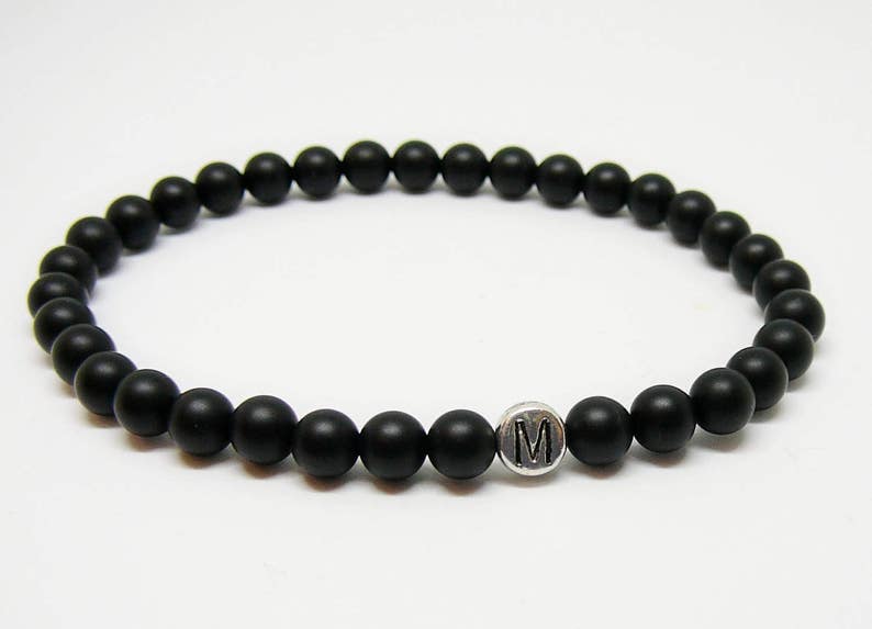 May include: Black beaded bracelet with a silver charm featuring the letter "M". The bracelet is made of small, round, matte black beads strung together. A simple, elegant piece of jewelry.