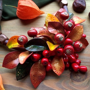 Puede incluir: Un broche hecho a mano de arcilla polimérica con un racimo de bayas rojas y hojas en tonos naranja, amarillo y verde. Las hojas tienen un brillo metálico y están acentuadas con purpurina dorada.