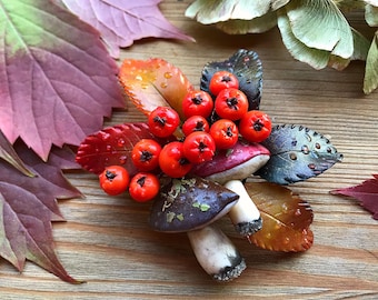 May include: A handmade brooch featuring a cluster of red berries, two mushrooms, and autumn leaves. The mushrooms are brown and white with a textured surface. The leaves are brown, orange, and green.