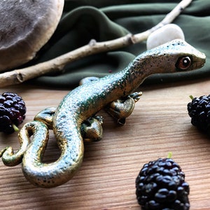 May include: A green and gold lizard figurine with a detailed textured surface. The lizard is posed with its head turned to the side and its tail curled around its body.