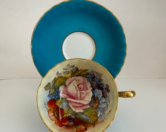 May include: A vintage teacup and saucer set with a floral design. The teacup features a hand-painted bouquet of pink, red, and blue flowers on a cream background. The saucer is a vibrant teal blue with a gold rim.