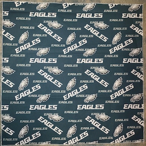 May include: A green fabric with a repeating pattern of white "EAGLES" text and a stylised eagle logo. The eagle is white with a black beak and black eyes.