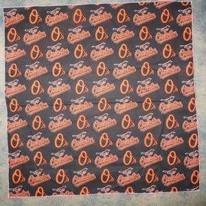 Op de afbeelding: Een zwarte bandana met een herhalend patroon van oranje "O's" en het woord "Orioles" in een gestileerd lettertype.