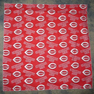 May include: A square fabric bandana featuring a repeating pattern of the Cincinnati Reds baseball team logo and the text Cincinnati Reds printed in white on a bright red background. The design is oriented in multiple directions across the cloth.