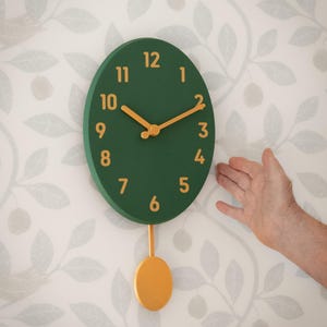 May include: An oval wall clock with a dark green face and gold-coloured numbers from one to twelve. The clock features gold-coloured hour and minute hands and a gold-coloured pendulum hanging from the bottom centre of the oval clock face.