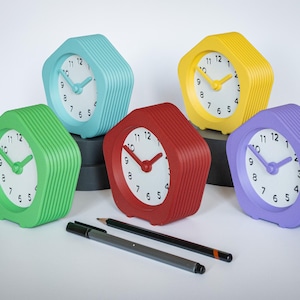 May include: Five hexagonal alarm clocks in green, blue, yellow, red, and purple. Each clock has a white face with black numbers and matching coloured hands. Two pens and a pencil are in the foreground.