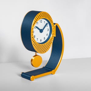 May include: A table clock with a dark blue circular frame and a yellow lattice border. The clock face is white with black numerals and dark blue hands. A yellow pendulum hangs from the clock, which is supported by a curved dark blue and yellow stand.