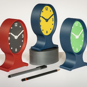 May include: Three oval-shaped table clocks with stands. One clock has a red frame and black face, one has a blue frame and yellow face, and one has a blue frame and green face. Each clock features white hands and numerals at 12, 3, 6, and 9.