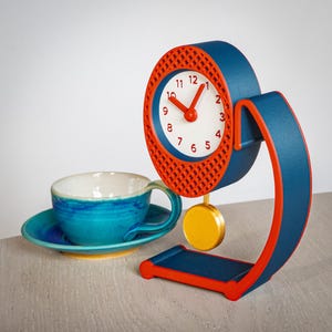 May include: A blue and orange desk clock with a white face, red numbers, and a gold-coloured pendulum, positioned next to a blue and white ceramic teacup and saucer. The clock features a circular frame with a textured orange lattice pattern.