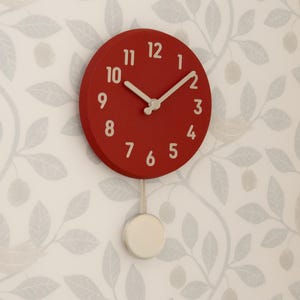 May include: A circular wall clock with a deep red face and light-coloured numerals from 1 to 12. The clock features matching light-coloured hour and minute hands and a pendulum with a circular weight hanging from the bottom of the clock face.