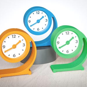 May include: Three analogue desk clocks with circular frames in orange, blue, and green. Each clock features a white face with black numerals at 3, 6, 9, and 12 o'clock, and matching coloured hands. The frames have a curved, open-base design.