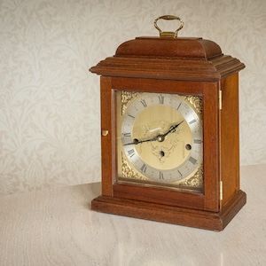 May include: A wooden mantel clock with a gold face and black Roman numerals. The clock has a handle and is set on a light surface. The clock face is visible through a glass panel. The clock measures approximately 20 cm in height.