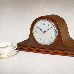 May include: A wooden mantel clock with an arched top, a white circular face, black numerals, and thin black hands. Beside the clock sits a small cream-coloured ceramic teacup and matching saucer with a thin blue and gold rim detail.