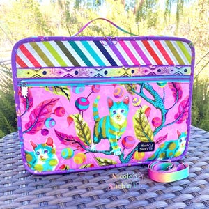 May include: Purple and pink laptop case with a colorful striped fabric on the top flap and a pink fabric with a cat and ball pattern on the bottom. The case has a zipper closure and a handle. A rainbow strap is included.