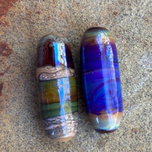 May include: Two cylindrical glass beads with smooth, polished surfaces. The left bead features horizontal bands of deep brown, olive green, and textured tan. The right bead displays a vibrant, iridescent swirl of royal blue, purple, and gold tones.