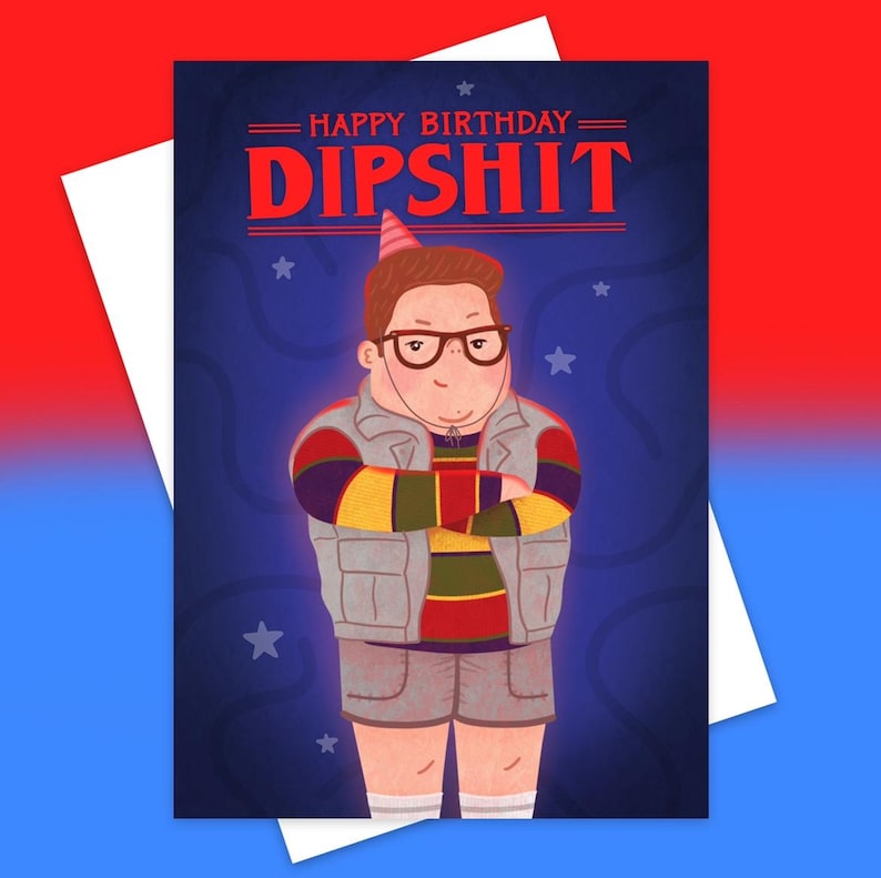 May include: Greeting card with a cartoon illustration of a person wearing glasses, a party hat, and a gray vest. The card reads "HAPPY BIRTHDAY DIPSHIT" in red letters against a blue background with stars. A white envelope is visible.