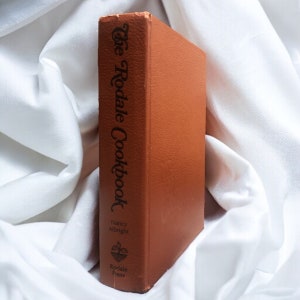 Puede incluir: Un libro de cocina encuadernado en cuero marrón titulado "The Rodale Cookbook" de Nancy Albright. El libro presenta un logotipo de planta verde con el texto "Rodale Press" debajo.