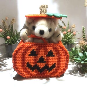 May include: A plush cream-colored teddy bear sits inside a handcrafted orange plastic canvas pumpkin container. The pumpkin features a black stitched jack-o'-lantern face, a brown stem, and a green leaf on top. A thin orange ribbon is tied around the bear.