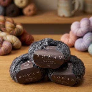 May include: Three skeins of dark grey textured yarn stacked together. Each skein features a brown paper label with the text ROWAN Soft Bouclé and 50g printed on the front. The yarn has a looped, curly appearance and is made of mohair, wool, and alpaca.