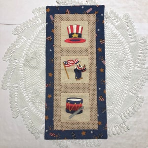 Op de afbeelding: Een blauwe en witte patriottische tafelrunner met een rood, wit en blauwe hoge hoed, een figuur die een vlag en een trommel vasthoudt, en een trommel met drumsticks. De runner is versierd met sterren en vuurwerk.