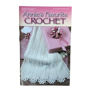 May include: A magazine cover for "Annie's Favorite Crochet" with a pink and white color scheme. The cover features a crocheted white blanket draped over a pink gift box with a bow. The magazine title is in large, decorative letters.