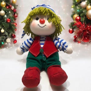 May include: A plush toy elf with green hair, a red and white striped shirt, a red vest, green pants, and red shoes. The elf is sitting with a big smile on its face.