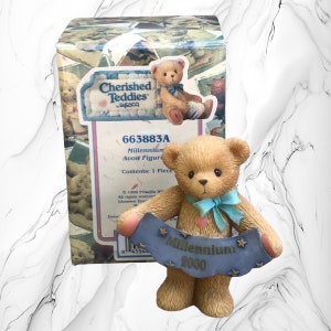 Puede incluir: Una figurita de oso de peluche marrón sosteniendo una pancarta azul con el texto "Millennium 2000" y estrellas. El oso lleva un lazo azul y tiene un corazón rosa en el pecho. La figurita está empaquetada en una caja con el texto "Cherished Teddies" y "663883A".