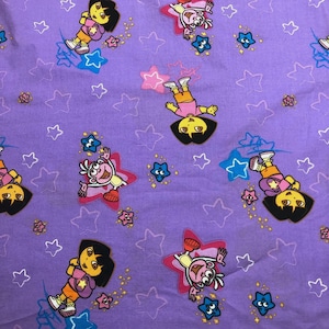 May include: Lilac fabric with a repeating pattern of Dora the Explorer characters and star shapes in various colours. The cartoon characters are in action poses, surrounded by smaller star accents.