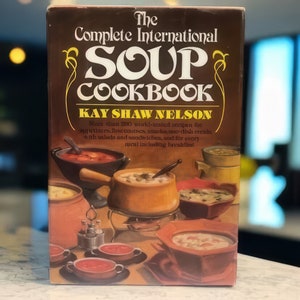 Può includere: Un libro di cucina vintage intitolato "The Complete International Soup Cookbook" di Kay Shaw Nelson. La copertina è marrone con scritte dorate e illustrazioni di varie zuppiere. Il libro promette ricette per antipasti, piatti principali, contorni e pasti.