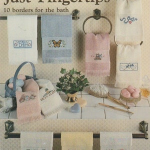 May include: A collection of embroidered bath towels in various colors, including white, blue, and pink. Each towel features a different cross-stitch design, such as butterflies, bears, and floral patterns. The image also includes a basket, soap, and a decorative brush.