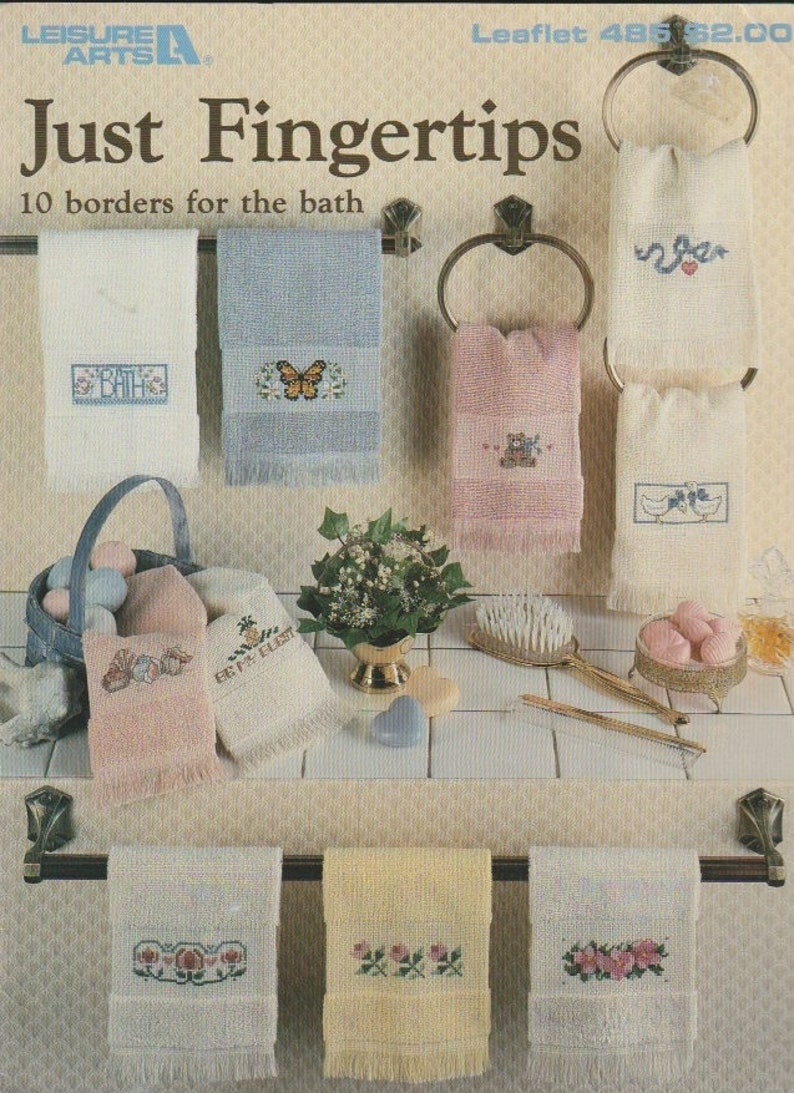 May include: A collection of embroidered bath towels in various colors, including white, blue, and pink. Each towel features a different cross-stitch design, such as butterflies, bears, and floral patterns. The image also includes a basket, soap, and a decorative brush.