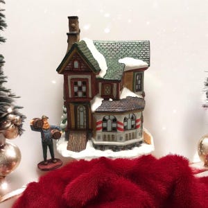 May include: A detailed Christmas village house with a green roof, snow accents, and a small figure carrying barrels. The house features brown and white paint, with a porch and chimney. The scene is set on a red plush surface.
