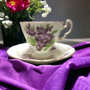 Op de afbeelding: Een witte kop en schotel set met een paarse viooltjes bloemen design en gouden rand. De kop en schotel staan op een paarse doek. Een vaas met bloemen staat op de achtergrond.