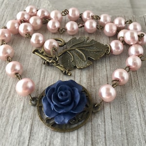 May include: A delicate necklace with a blue rose charm and a leaf pendant. The necklace is made with pink pearls and a bronze chain.