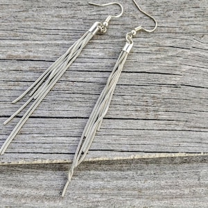 May include: Silver-toned dangle earrings with multiple strands of delicate chains. The earrings have a simple hook design and a small cylindrical cap where the chains are attached. The background is a weathered wooden surface.