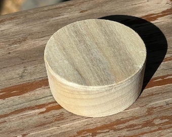 May include: A round, unfinished wooden object with a light, natural wood grain. The object is cylindrical, with a flat top and bottom, and appears to be made of stacked layers of wood. The object is sitting on a weathered wooden surface.