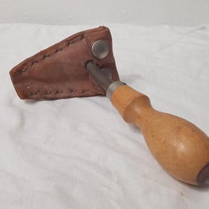 May include: A woodworking chisel with a wooden handle and a metal blade, partially encased in a brown leather sheath. The sheath is secured with a metal snap. The chisel is designed for shaping and carving wood.