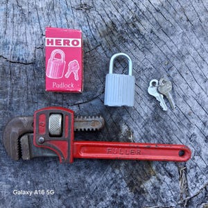 May include: A red and black pipe wrench with the word "FULLER" on the handle, a gray padlock with keys, and a box of "HERO" padlocks with keys. The items are on a weathered wood surface.