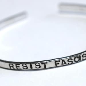 May include: A silver-colored cuff bracelet with the words "RESIST FASCISM" stamped on the front. The bracelet is open-ended and appears to be made of a thin metal band.
