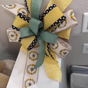 May include: A decorative bow made from yellow, green, and cream ribbons. Some ribbons feature a bee and sunflower design, while others have black polka dots. The bow is likely intended for decorative purposes.